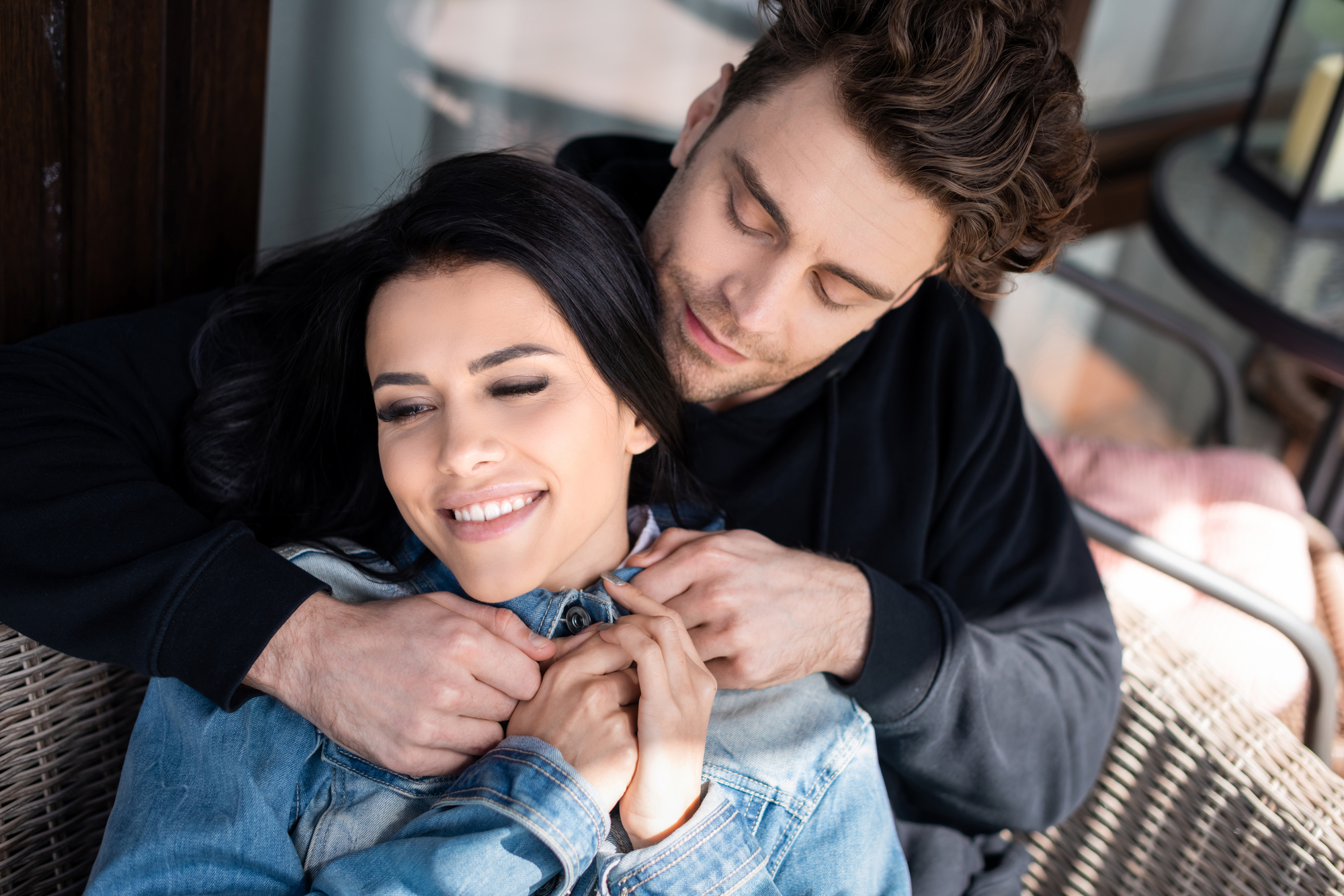 High angle view of man hugging smiling girlfriend on wicker sofa on terrace